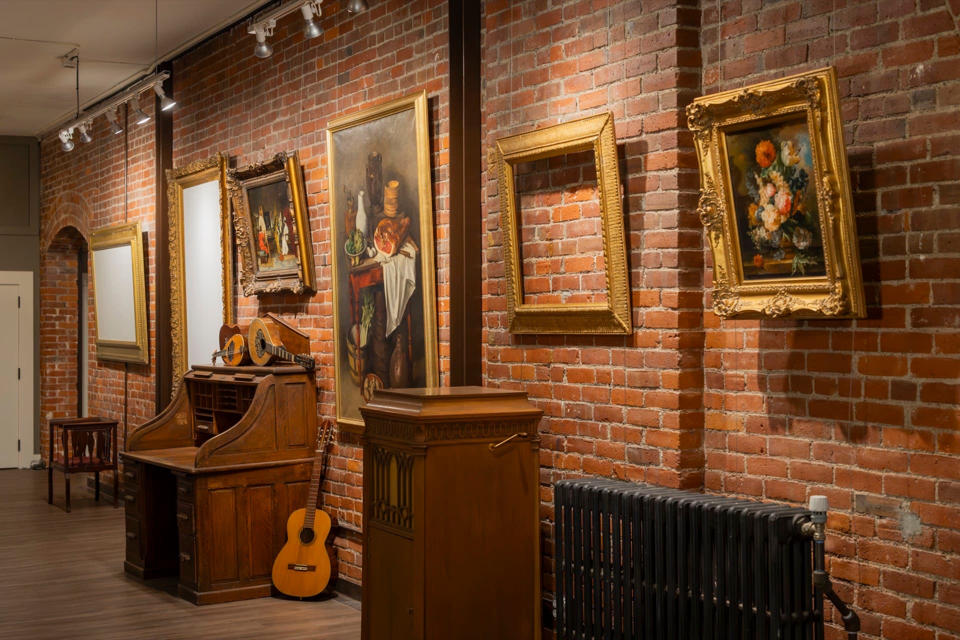 Gilt-framed paintings on the exposed brick wall of the gallery, an antique desk and a guitar below